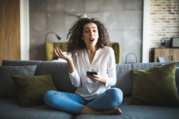 Portrait of a suspicious woman holding phone looking at you sitting on a sofa in the living room of...