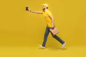 Fototapeta na wymiar Portrait of young delivery man in yellow uniform with paper packet and takeaway coffee isolated over yellow background.