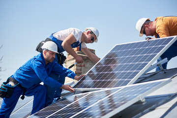 Male team engineers installing stand-alone solar photovoltaic panel system. Electricians mounting blue solar module on roof of modern house. Alternative energy concept.