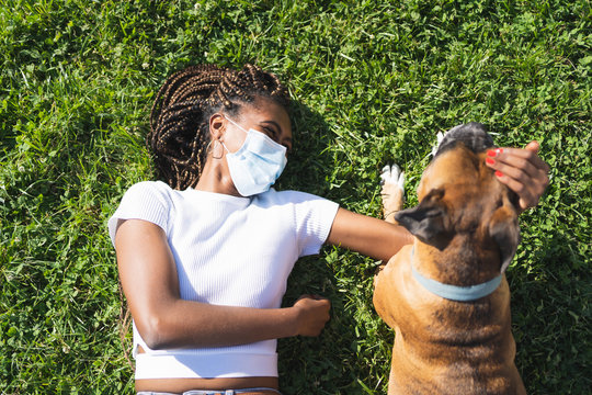 Woman with a protective mask stretched out on the grass and stroking the head of a boxer dog