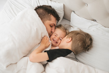 Young caucasian father plays with children in bed weekend. Cuddle daughter and son in the morning. Close up family portrait. Father's day concept