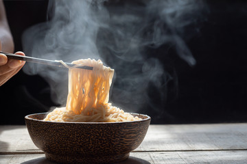 Hand uses chopsticks to pickup tasty noodles with steam and smoke in bowl on wooden background,...