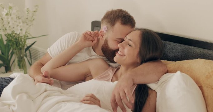 Man and woman in romantic relationship cuddling and smiling while lying on bed in morning. Couple in love embracing and spending time together in bedroom Concept of leisureand family.