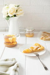 Piece of homemade shortbread lemon pie sprinkled with powdered sugar on a plate. Metal fork for a dessert, tea in a mug. White flowers in a jug, honey in a jar and lemon slices in the background.