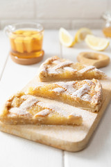 Three pieces of homemade shortbread lemon pie sprinkled with powdered sugar on a cutting board. Tea in a mug, honey and lemon slices in the background. Close-up