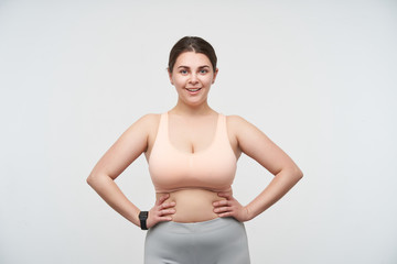 Fototapeta na wymiar Indoor photo of brunette chubby female with ponytail hairstyle dressed in sporty clothes while standing over white background, keeping her hands on waist and smiling cheerfully at camera