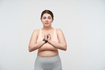 Fototapeta na wymiar Studio shot of young lovely dark haired oversized woman with casual hairstyle keeping folded hands on her chest and looking hopefully at camera, posing over white background