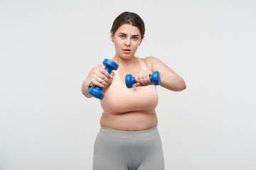 Studio shot of young plump oversized woman with ponytail hairstyle keeping dumbbells in raised hands while making fitness at home, isolated over white background