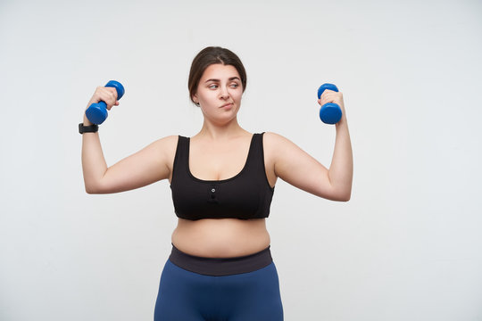 Bewildered young dark haired plump female with casual hairstyle raising hands with dumbbells and looking confusedly at it, isolated over white background. Concept of weight loss