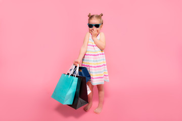 Little blonde girl wearing sunglasses with many shopping bags on a pink studio background.