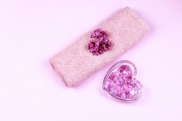 Heart shaped glass plate and rolled towel with lilac flowers on light pink background. Beautiful spa composition plat lay.