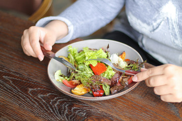 Woman is eating healthy vegetables and beef salad with knife and fork on wooden table
