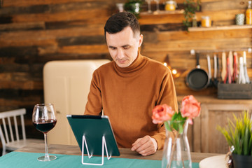 Young man in casual clothes using digital tablet, online communication, internet surfing, high glass with red wine on the table.