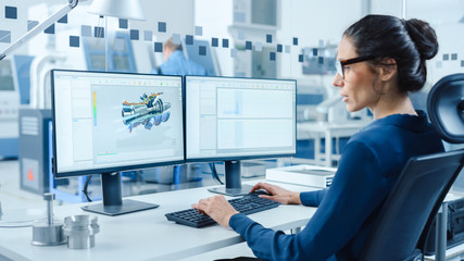 Industrial Female Engineer Working on a Personal Computer, Two Monitor Screens Show CAD Software with 3D Prototype of Engine and Charts. Modern Factory with High-Tech Machinery - obrazy, fototapety, plakaty