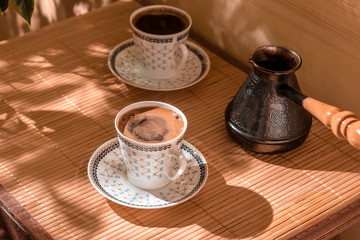 Two cups of coffee and cezve on the table in the patio lit by morning sun.