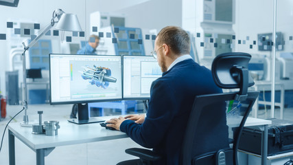 Industrial Engineer Working on a Personal Computer, Two Monitor Screens Show CAD Software with 3D Prototype of Hybrid Electric Engine and Charts. Working Modern Factory.
