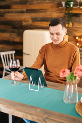 Young happy man communicates on digital tablet, sitting at table in the bright kitchen room, holding tall glass of red wine in his hand.