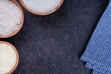 Dark marble background with rice flour, corn flour and whole grain flour in wooden bowls and blue napkin. Baking ingredients with space for text or image. Flat lay top view, studio shot