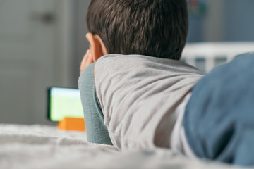 back view of boy watching online lesson on smartphone while lying on bed a home