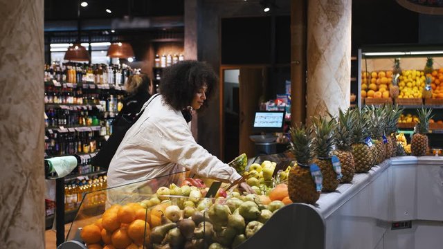 Afro american girl in casual outfit, with string bag and headphones on neck. She smiling, selecting apples and pears at fruit department of a shop