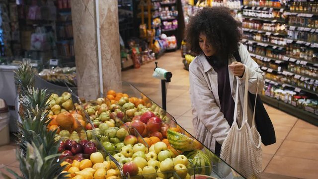 Afro american lady in casual clothes, with headphones on neck. She smiling, selecting apples, putting into string bag at fruit department of grocery
