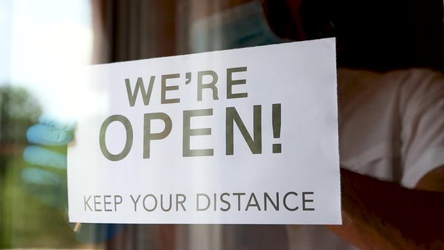 Business owner with gloves and face mask hanging an open sign "We're open. Keep your distance" at a restaurant. Concept of new normality due to coronavirus