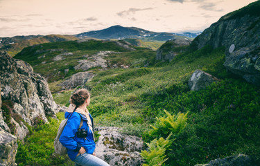 Young woman traveler in a blue jacket with a backpack and a photo camera in the mountains, against the backdrop of a beautiful landscape