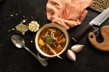 Vegetable soup with lamb, onions, garlic and potatoes. Soup plate, spoon, knife, towel and ingredients on a black table. Top view.