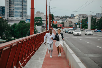 Young interracial couple runs on bridge and laughs cheerfully on the background of the city and the road.