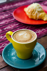 A cup of latte is served with colorful ceramic cups, side by side with food and cutlery. Everything is photographed on the table with traditional woven cloth ornaments ..