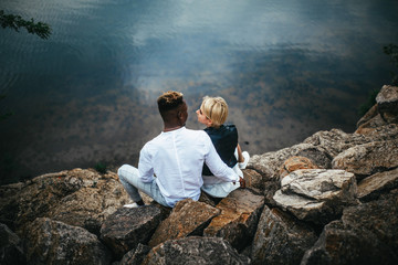 Interracial couple sits on rocks and hugs against background of river.