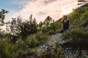 A full body shot of a relaxed young Caucasian female hiker sitting on the ground on a hiking path in the French Alps during sunset