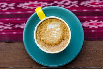 A cup of latte is served with colorful ceramic cups and photographed on the table with traditional woven cloth ornaments.