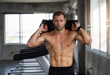 Fototapeta na wymiar Muscular young man exercising with a heavy sandbag in a fitness gym. Sporty man working out with heavy sandbag
