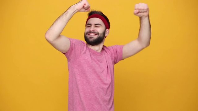 Young bearded fun fitness sporty guy 20s sportsman in headband pink t-shirt spend weekend in home gym dancing fooling around isolated on yellow background. Workout sport motivation lifestyle concept