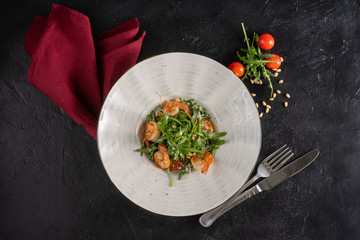 Fresh green salad with fried shrimp, cherry tomatoes and arugula in a deep plate on a black background. Top view.