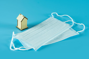Creative concept, medical mask with tiny wooden house isolated on blue background, representing to motivate people to stay at home, recommendation to prevent spreading coronavirus.