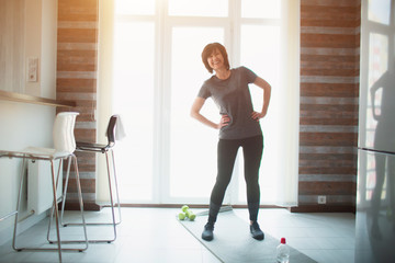 Adult fit slim woman has workout at home. Cheerful positive senior female person hold yoga mat in hand and point on it. Smiling during standing in room before workout.