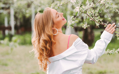 Back view young beautiful gentle woman with wavy hair in white shirt posing with blossom tree on countryside garden. Happy carefree romantic hippie girl relax and enjoy spring nature in green park