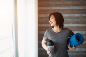 Adult fit slim woman has workout at home. Cheerful positive senior female person smiling and looking at window. Woman after exercising drink protein shake and hold yoga mat.