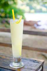 Isolate of a glass cold lemon smoothie juice place on the wooden table with brown color. Blur backdrop is sea, sky, beach, and trees. Feeling refreshing and Sour taste. Summer and vacation concept.