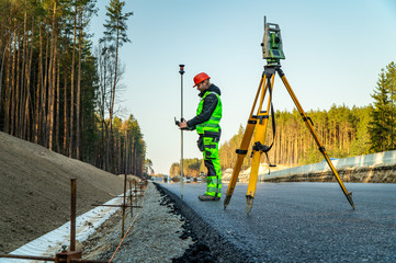 Surveyor engineer with equipment (theodolite or total positioning station) on the construction site of the road or building with construction machinery background - obrazy, fototapety, plakaty