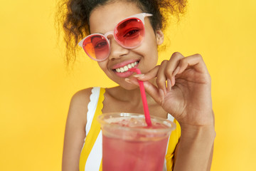 Smiling pretty african teen gen z girl holding straw drinking ice drink in hands wear pink sunglasses stylish glasses enjoy cool cocktail party isolated on yellow summer studio background. Portrait