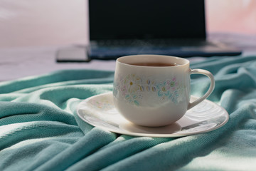 A cup of tea on the bed and light blue blanket with sunrise, Morning teatime with notebook computer and smart phone, Normal practice by working at home during covid-19 outbreak around the world.
