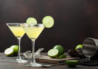 Gimlet Kamikaze cocktail in martini glasses with lime slice and ice on wooden board with fresh limes and strainer with shaker.