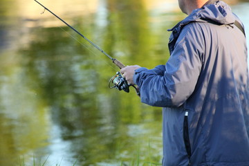 A fisherman in a jacket with a spinning rod in his hand winds a fishing line on a reel close-up on blurred water with shore trees reflection background, fishing on the river on a summer day