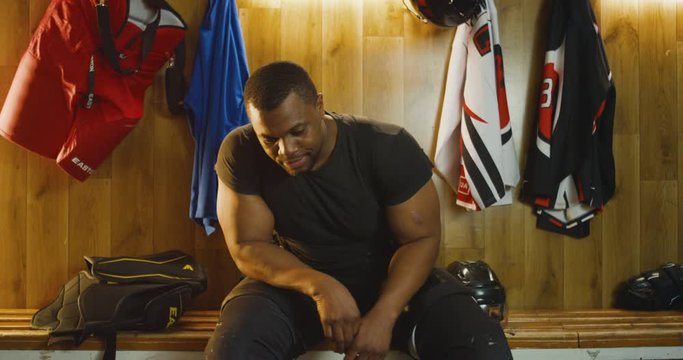 Portrait of young handsome and strong African American male hockey player sitting on bench in dressing room, taking off uniform and smiling cheerfully to camera. After game in change room.