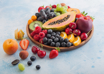 Fresh raw organic summer berries and exotic fruits in round wooden plate on light blue kitchen background. Papaya, grapes, nectarine, orange, raspberry, kiwi, strawberry, lychees, cherry and physalis.