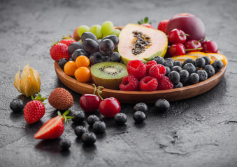 Fresh raw organic summer berries and exotic fruits in round wooden plate on black kitchen background. Papaya, grapes, nectarine, orange, raspberry, kiwi, strawberry, lychees, cherry and physalis.