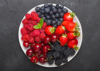Fresh organic summer berries mix in white plate on black kitchen table background. Raspberries, strawberries, blueberries, blackberries and cherries. Space for text